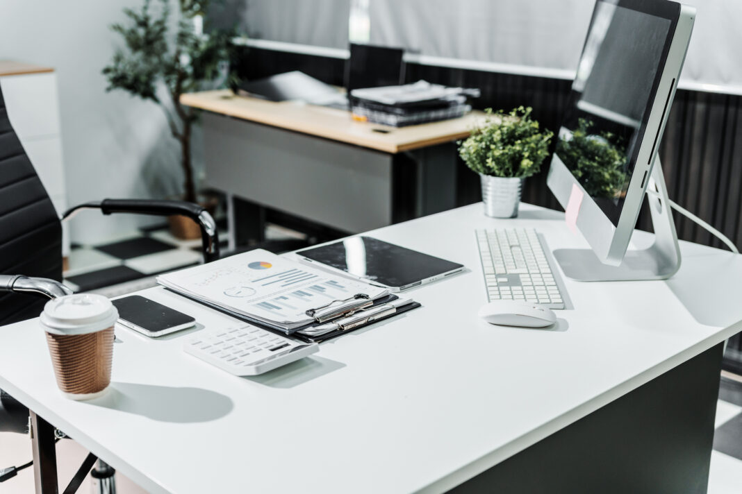 A modern, white-colored business office with a wooden desk, calculator, desktop computer, and a black leather chair. The executive office features private office furniture for the chief of staff.