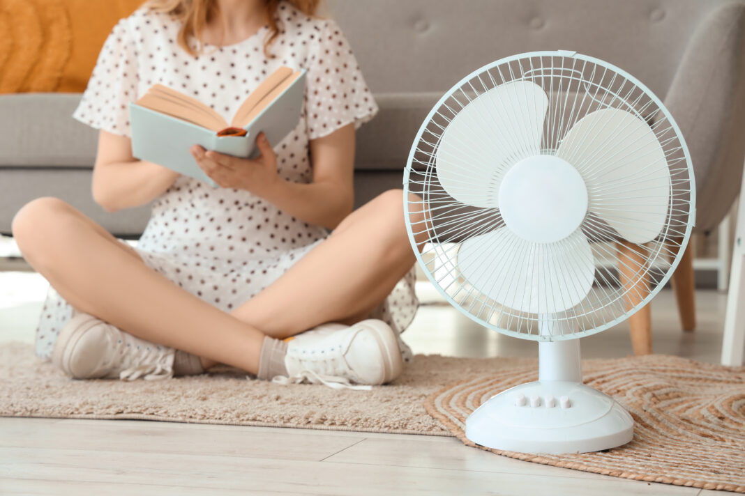 Young woman with electric fan sitting on floor and reading book in living room