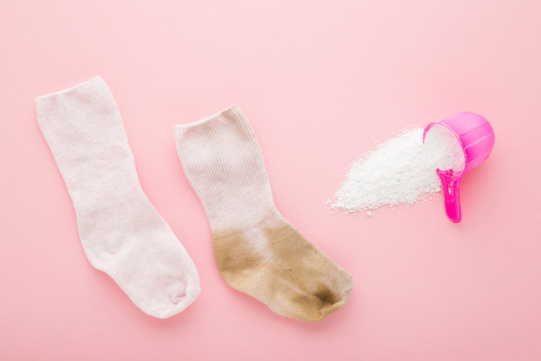 Baby socks and cup or scoop of white powder on light pink table background. Pastel color. Closeup. Detergent for clothes washing. Problem and solution. Compare dirty and clean socks. Top down view.